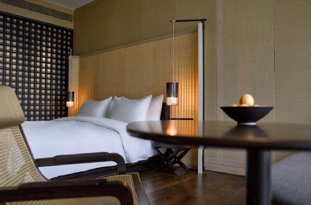 The Ruma Hotel And Residences Kuala Lumpur Eksteriør billede The photo shows a well-designed bedroom with a modern and minimalist aesthetic. The room features a large bed with white linens, positioned against a textured wall. On either side of the bed, there are pendant lights that provide a warm glow. The dec