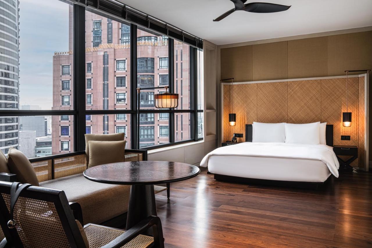 The Ruma Hotel And Residences Kuala Lumpur Eksteriør billede The photo shows a modern hotel room featuring a large bed with white linens, situated against a backdrop of large windows that allow for ample natural light. The room includes a wooden floor and has a minimalist aesthetic, with a round table and two 