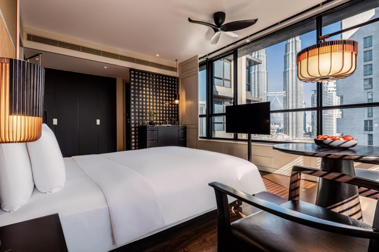 The Ruma Hotel And Residences Kuala Lumpur Eksteriør billede The photo shows a modern hotel room featuring a large bed with white linens. There's a ceiling fan above and large windows letting in natural light, providing a view of a cityscape outside. To one side, there's a small seating area with a round table